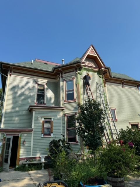 Man on ladder painting a two-story house with green siding, brown trim, and a decorative peak.