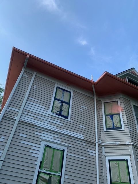 Exterior of a house with beige siding, red roof, and windows taped for painting against a blue sky.