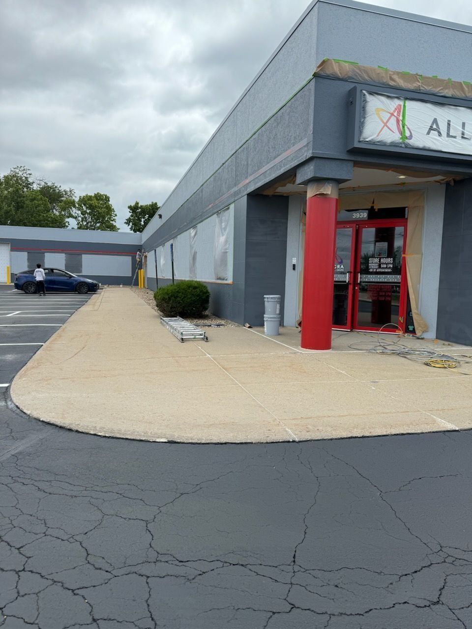 Exterior of a commercial building with a red pillar, gray facade, and cracked asphalt parking lot.