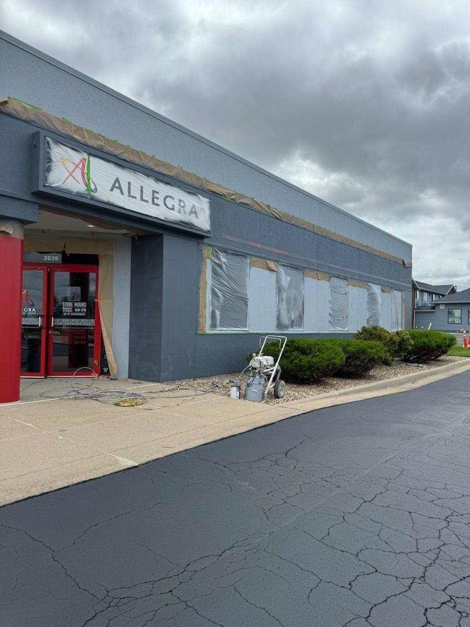 A gray Allegra storefront with a red door, cloudy sky, and construction underway.