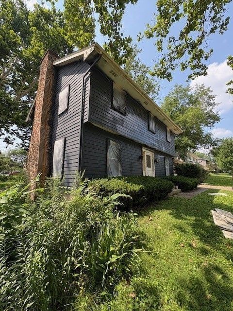 Dark blue two-story house with boarded windows, brick chimney, and overgrown landscaping.