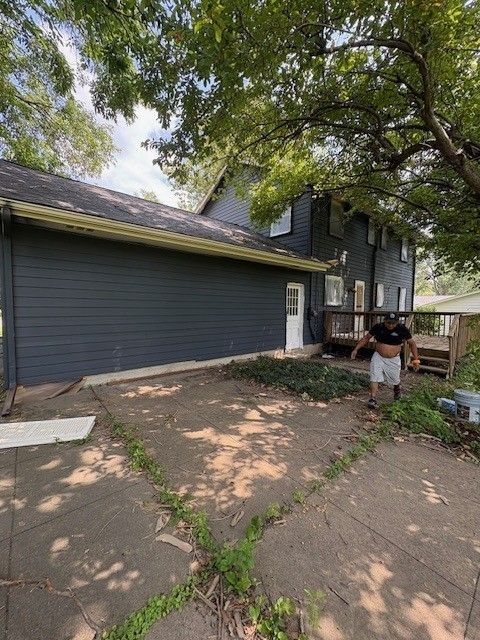 Person standing near dark blue house with concrete patio, overgrown with weeds.
