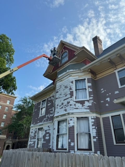 House being painted; a worker in a lift repairs the exterior. Purple siding, peeling paint, blue sky.