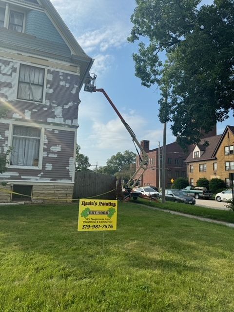 House being painted by a boom lift. Brown house with peeling paint, worker in the lift, green lawn, blue sky, and parked cars.