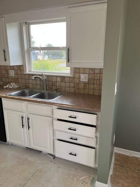 White kitchen cabinets with black handles, stainless steel sink, tiled backsplash, window.