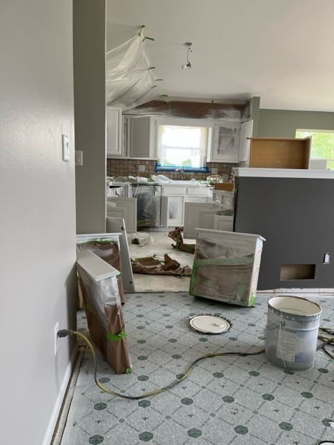 Kitchen undergoing renovation: cabinets, walls, and floor covered in plastic, paint cans visible.