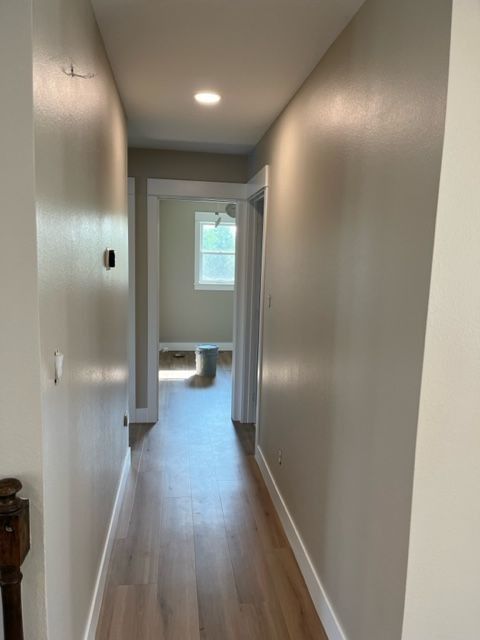 Narrow hallway with light wood floor, beige walls, and a bright room visible at the end.