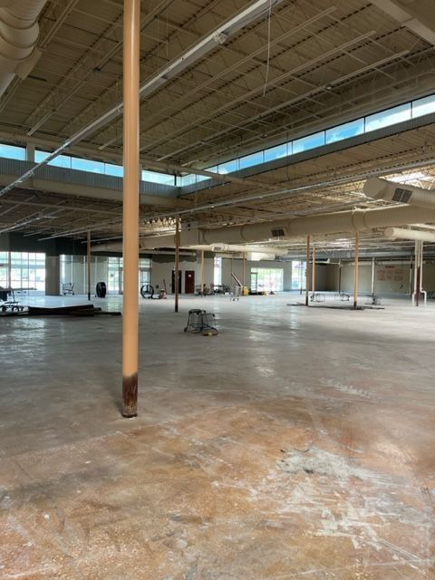 Empty, large commercial space; concrete floor, exposed beams, interior columns, high ceiling, natural light from windows.