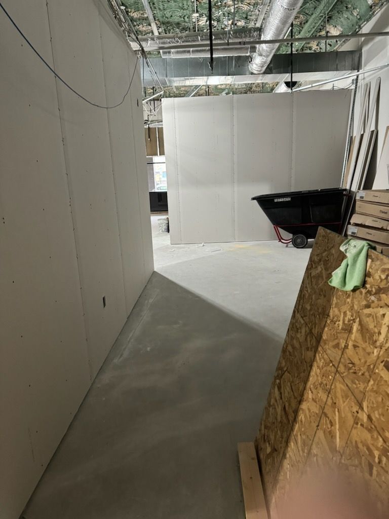 A narrow construction area with drywall walls and concrete floor; a trash container is in the distance.