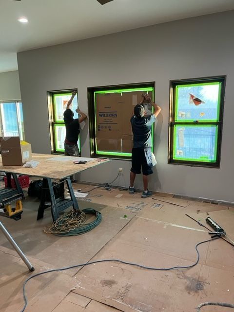 Two people installing windows in a room. Green-taped window frames, cardboard flooring, gray wall.
