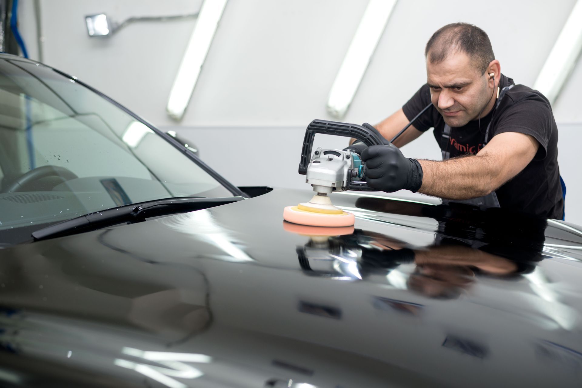 A professional using a power buffer to polish the hood of a black car in a brightly lit garage.