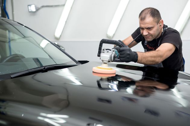 A professional using a power buffer to polish the hood of a black car in a brightly lit garage.