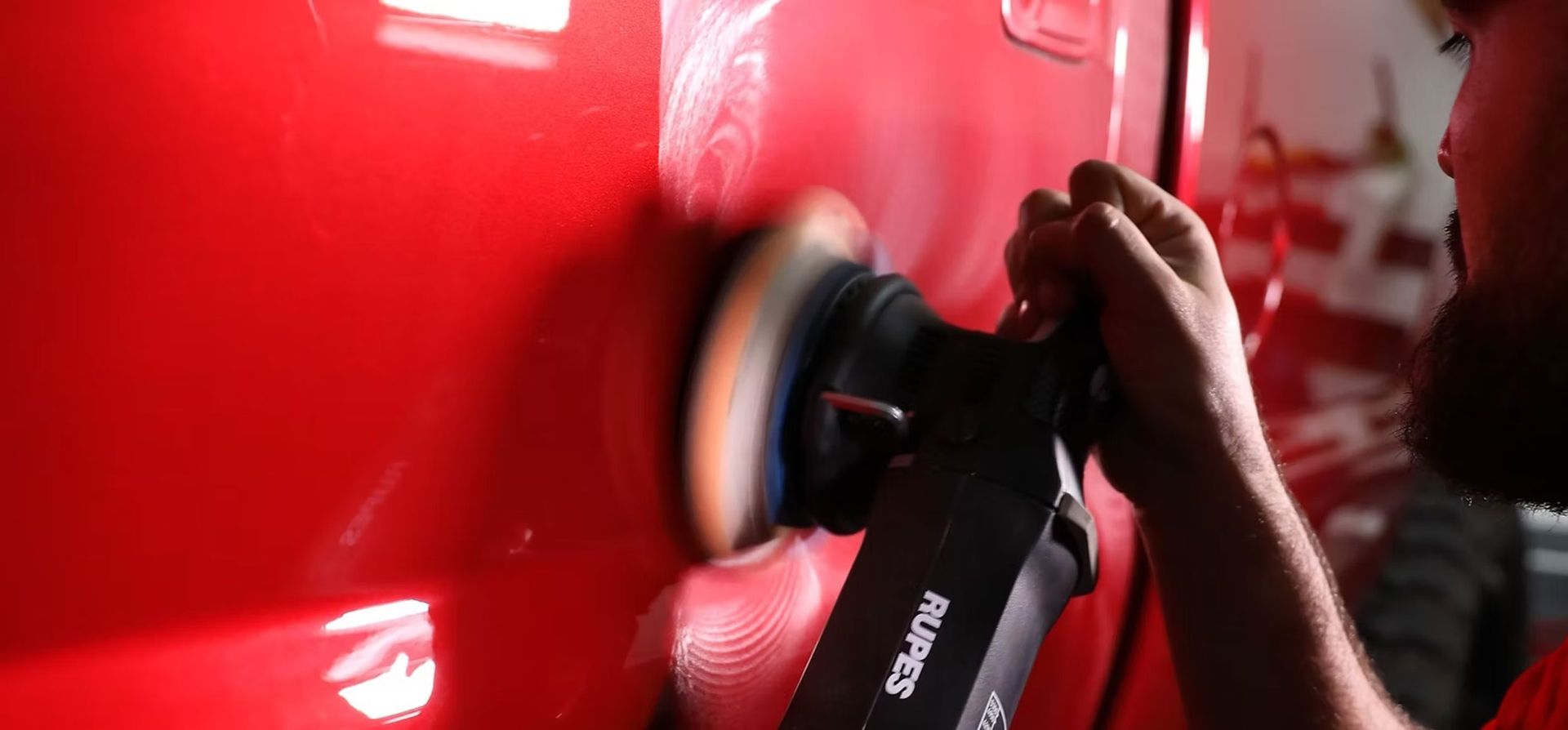 A person uses a RUPES power buffer to polish a bright red vehicle panel in a workshop.