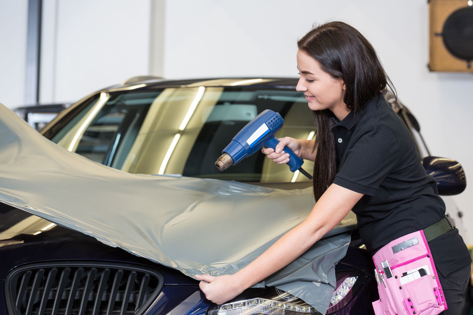 A technician uses a heat gun to apply a grey vinyl wrap to the hood of a car in a workshop.