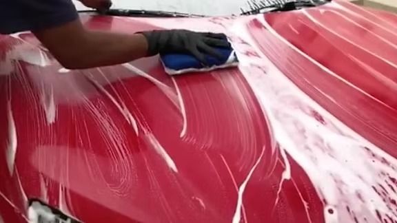 A person wearing a black glove uses a blue sponge to wash a red car hood covered in soap suds.
