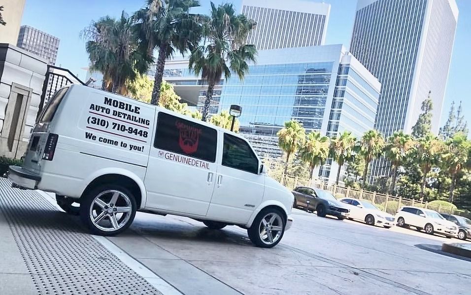 A white mobile auto detailing van parked on a city street, with modern skyscrapers and palm trees in the background.
