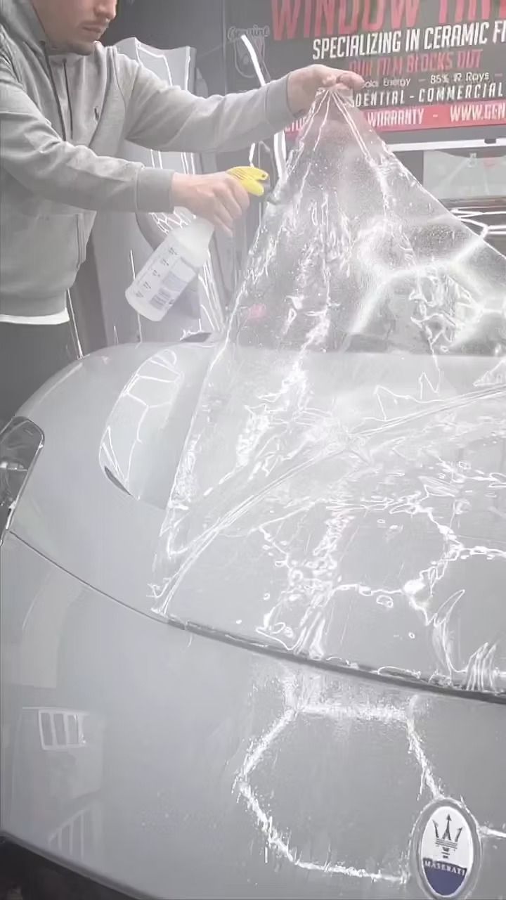 A person sprays water on a gray Maserati hood while applying a clear paint protection film.