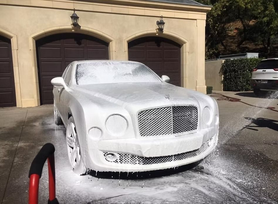 A Bentley covered in thick white cleaning foam, parked in a driveway in front of a garage.