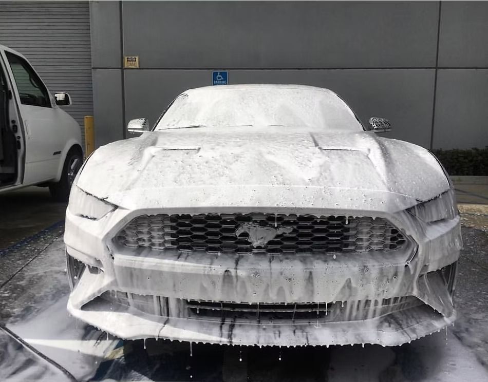 A Ford Mustang covered in thick white soap suds during a car wash in a parking lot.