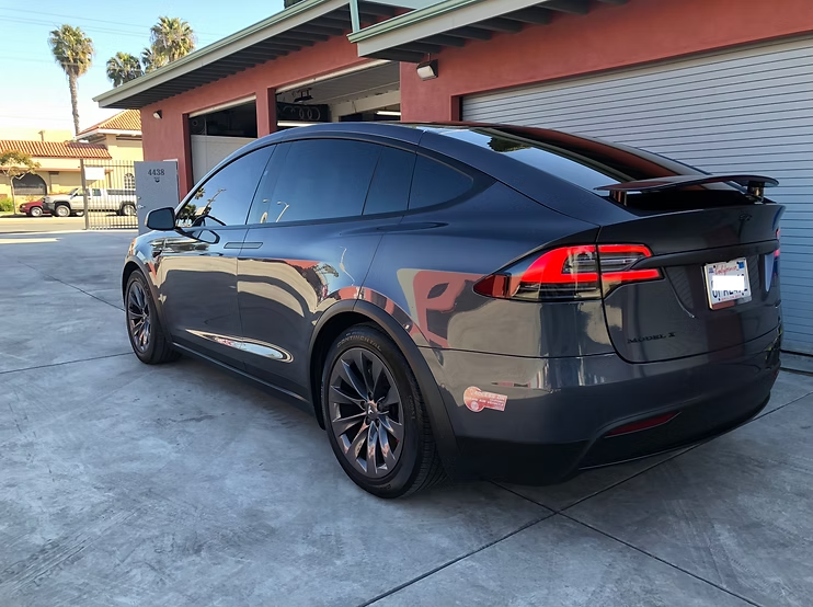 A dark gray Tesla Model X parked on a concrete driveway in front of a building with red exterior walls.