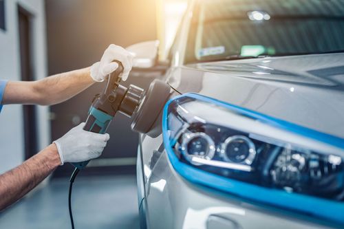 A professional wearing gloves uses an electric buffer to polish the side of a silver car with blue protective tape applied.