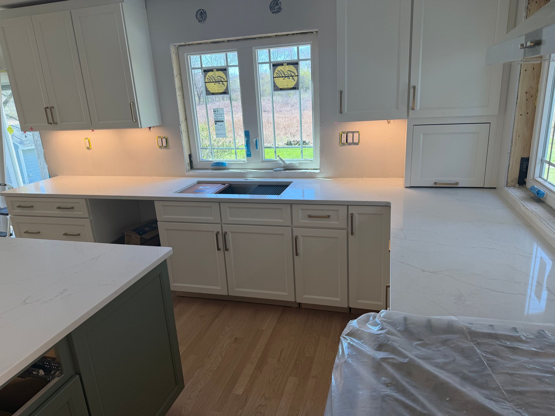 Bright white kitchen with gray cabinets, wood floor, central sink, and plastic-covered floor area.