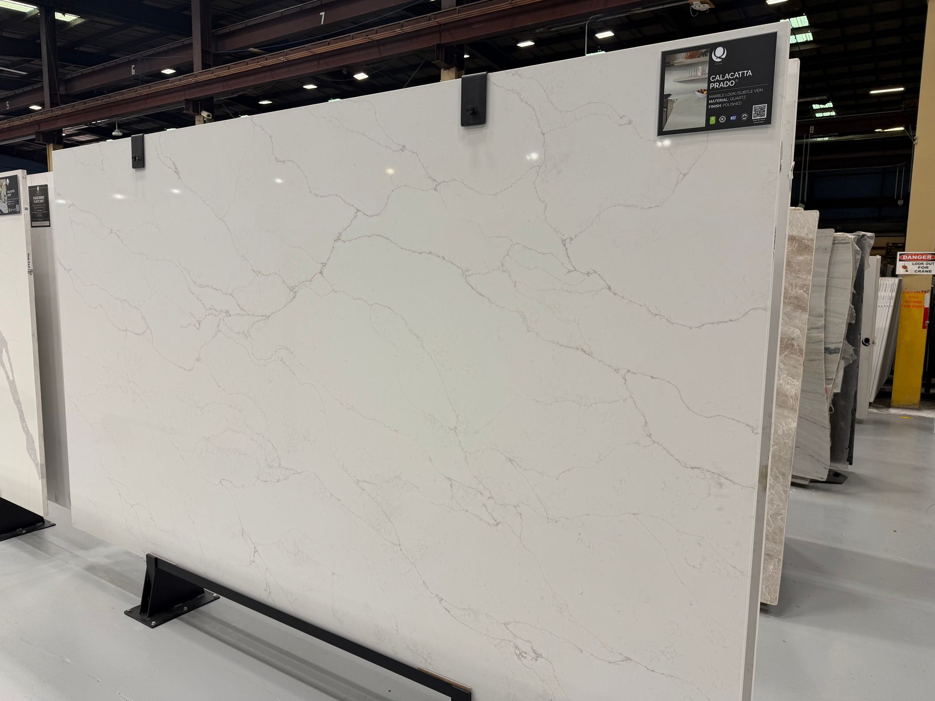 White marble-look slab display in a warehouse showroom, with subtle gray veining and a small corner label.