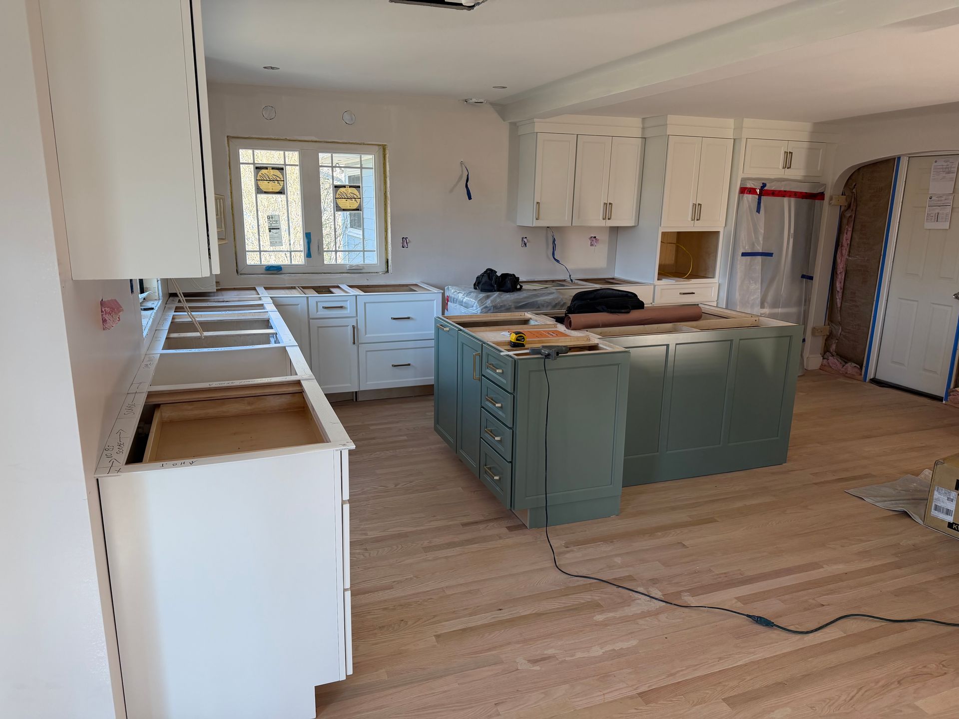 Unfinished kitchen with white cabinets, teal island, bare floor, and open windows.