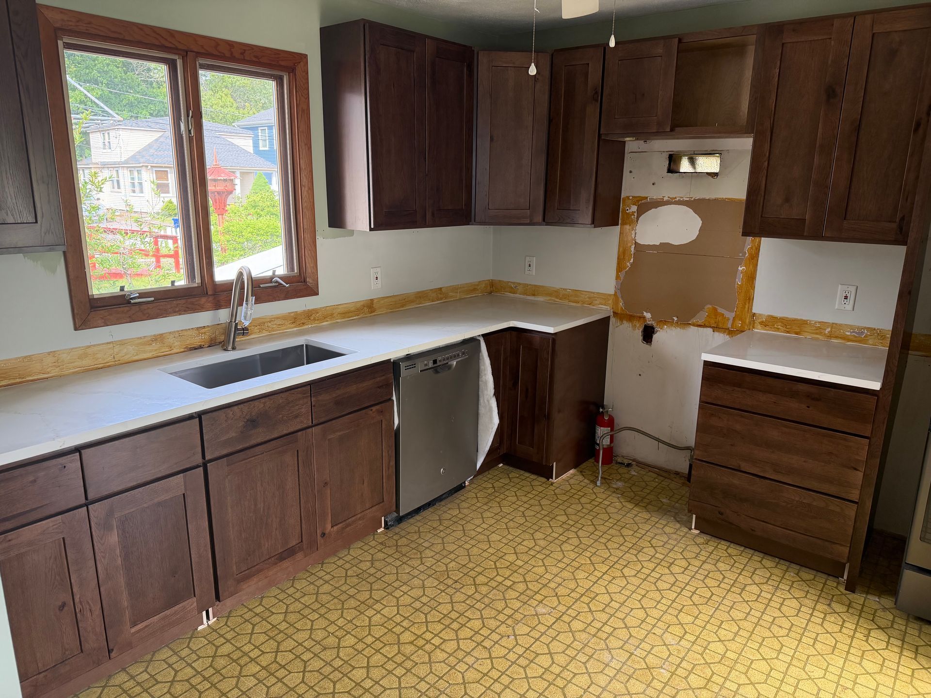 Empty kitchen with dark wood cabinets, white countertops, a window above the sink, and yellow patterned flooring.