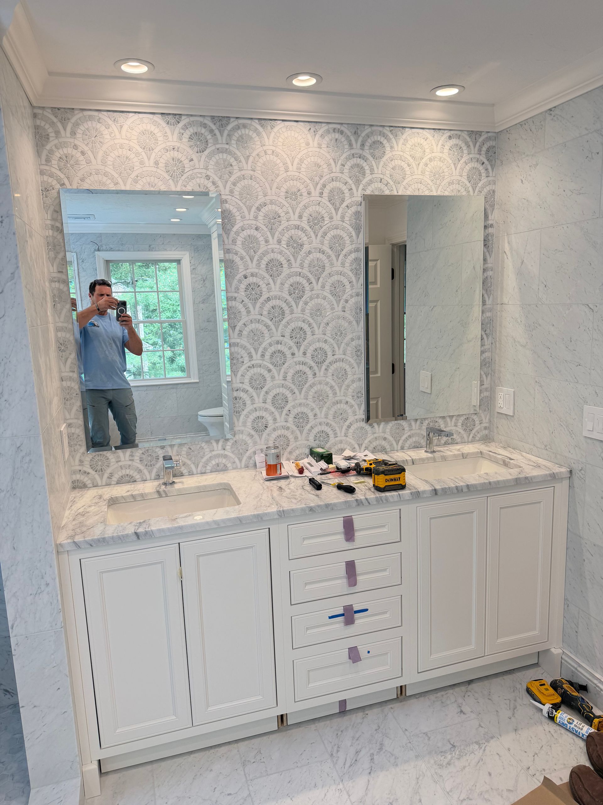Bathroom vanity with marble countertop, white cabinets, and patterned wallpaper; person reflected in mirror.