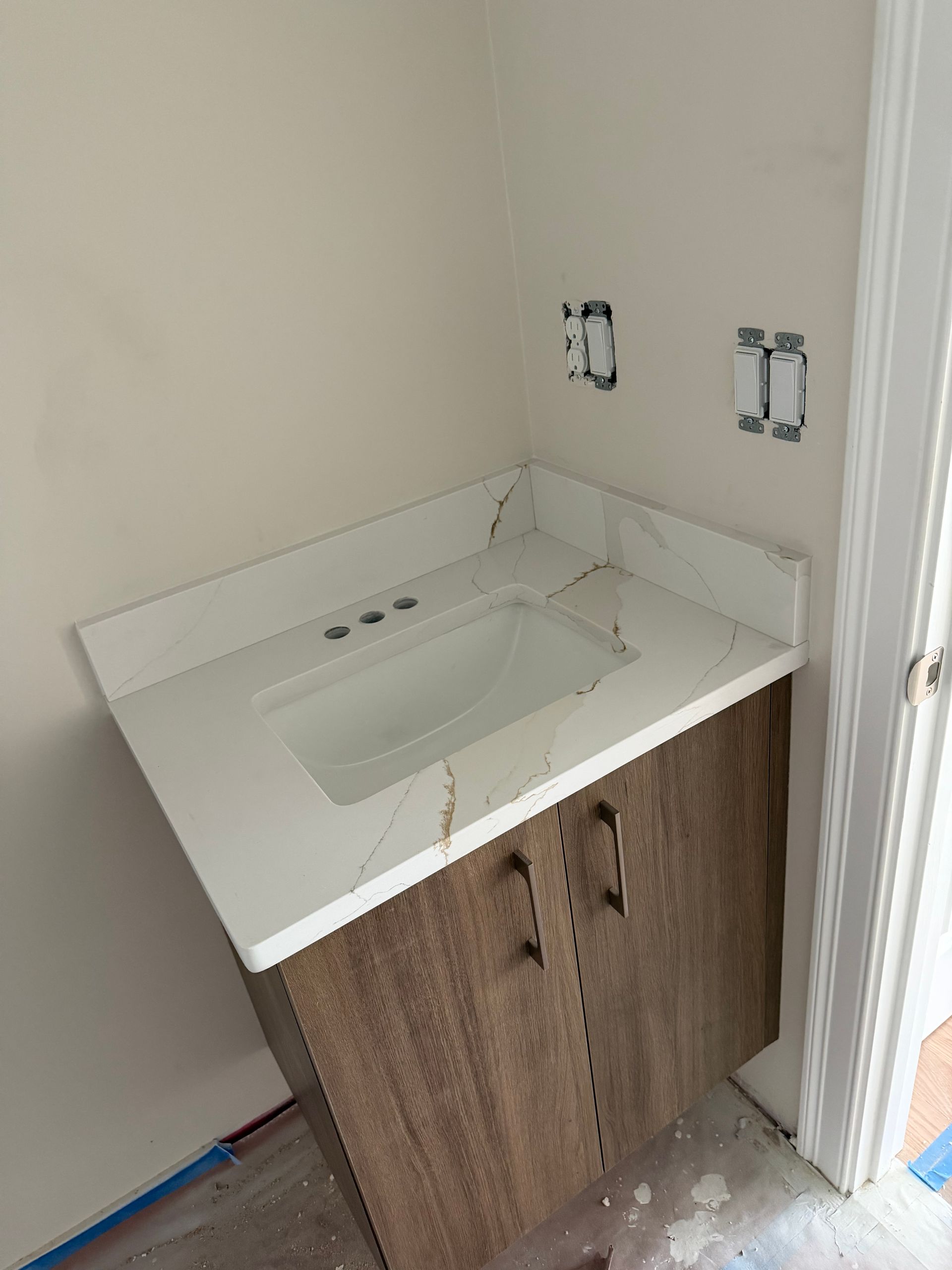 Bathroom vanity with white sink top and unfinished wood cabinet against a wall with exposed plumbing hookups