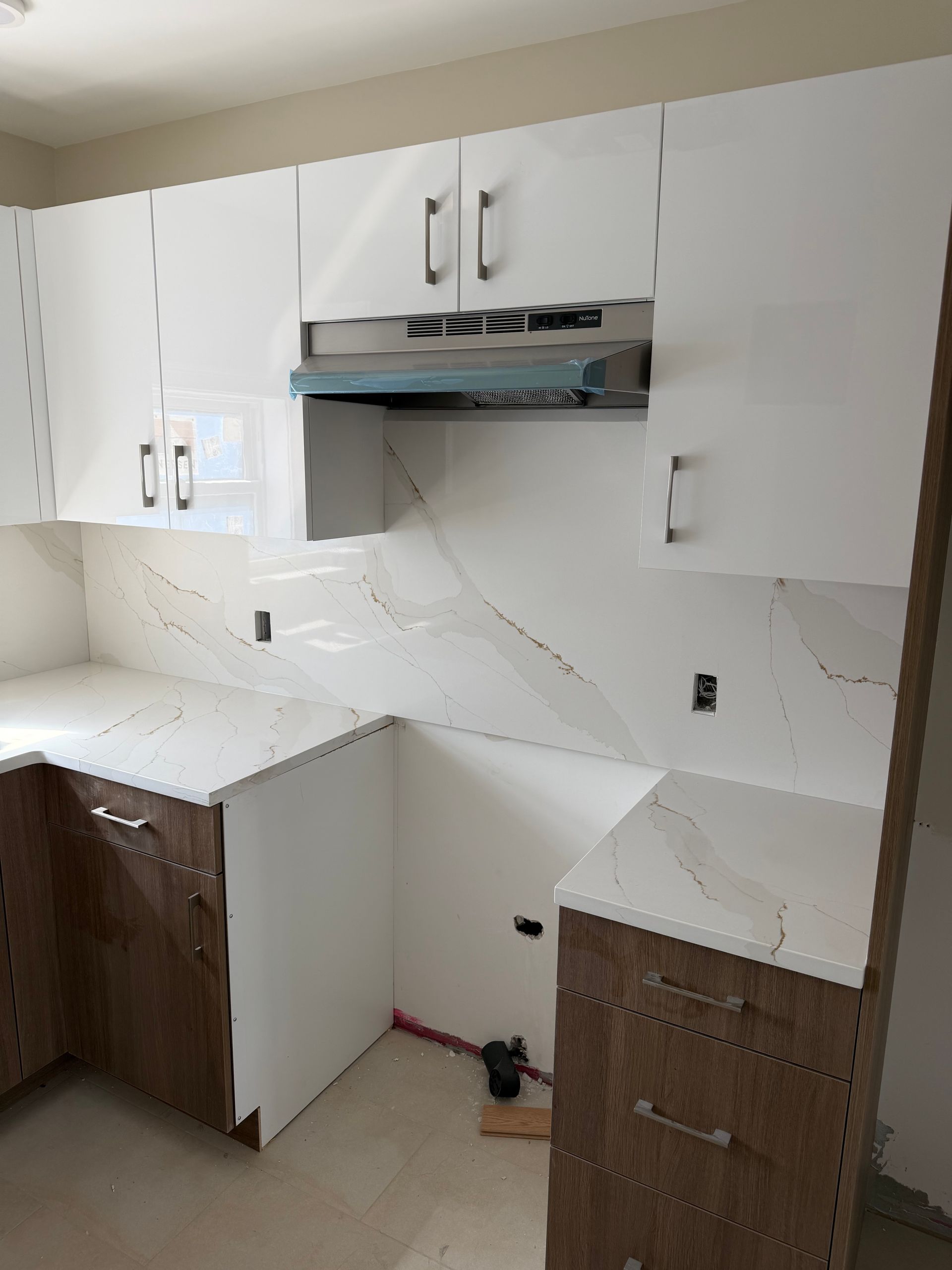 Small kitchen with white cabinets, marble-look counters, and a stainless range hood under construction.