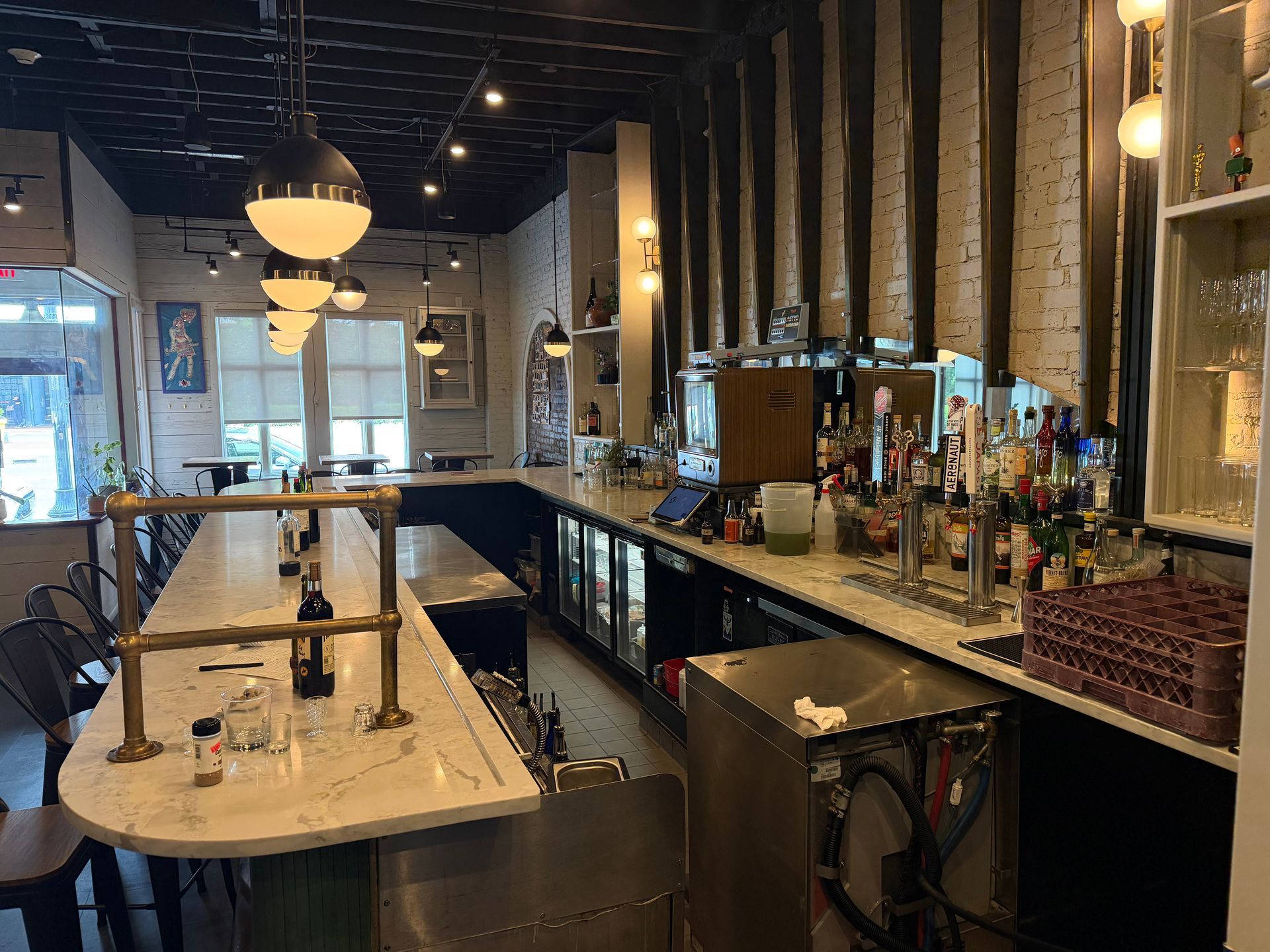 Empty bar interior with long counter, shelves of bottles, pendant lights, and exposed brick walls