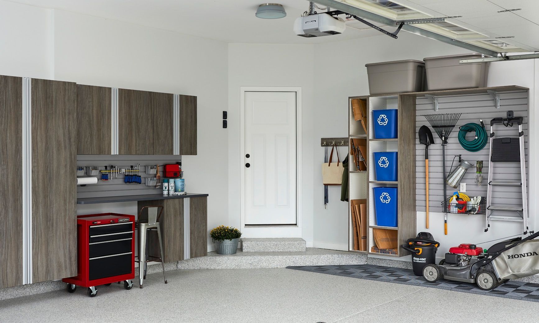 Cluttered garage with cabinets, workbench, tool chest, shelves, and lawn equipment