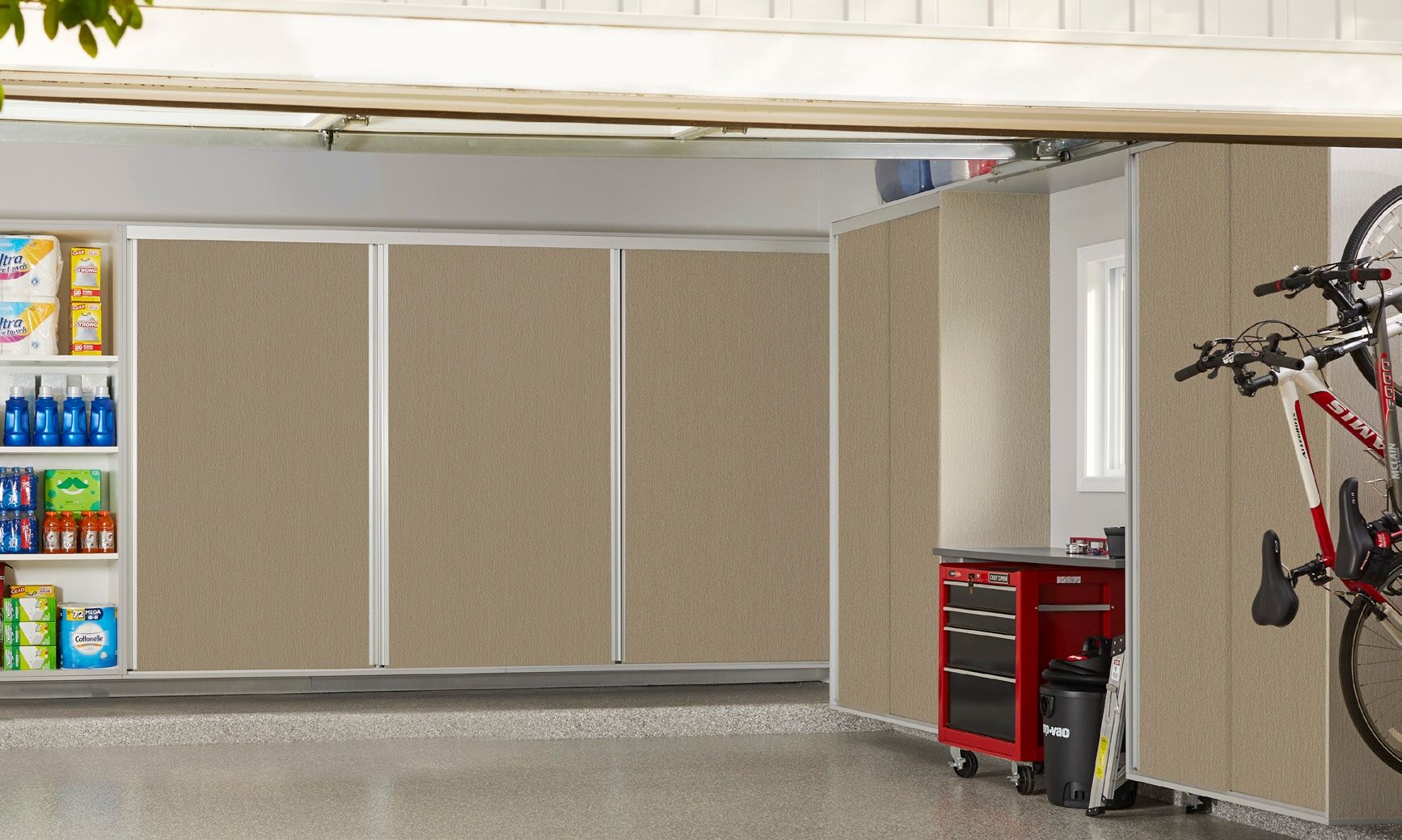 Garage with beige cabinet doors, shelving of supplies, and a red tool chest beside a bicycle on the right.