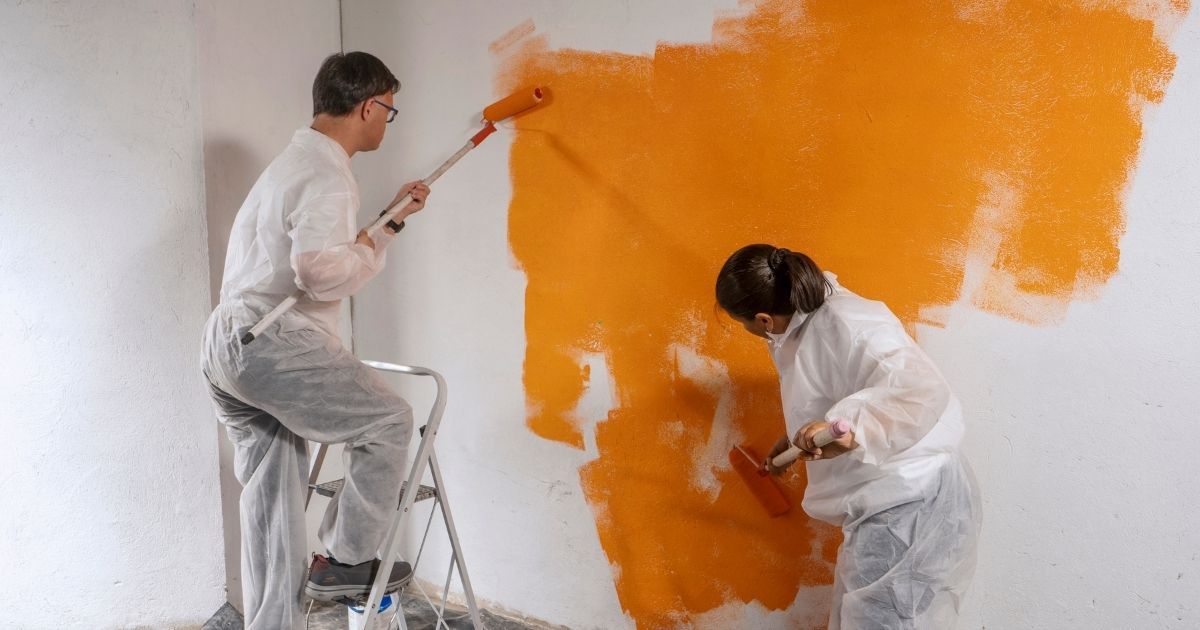 Two painters in white coveralls painting a white wall bright orange indoors