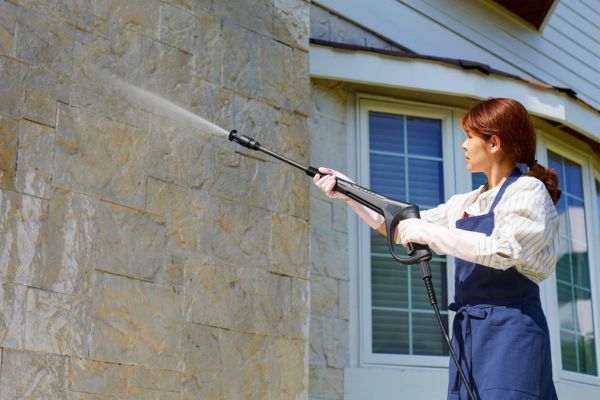 Woman power-washing a stone house exterior with a long spray wand