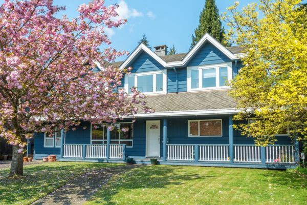 Blue house with white trim and porches, framed by blooming pink and yellow trees in a sunny yard
