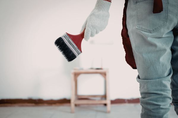 Gloved hand holding a red paintbrush beside a small stool in a room with white walls