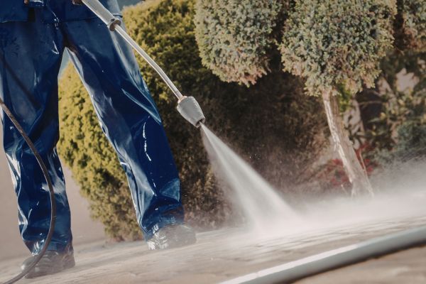 Person in blue pants pressure-washing a concrete surface outdoors near bushes