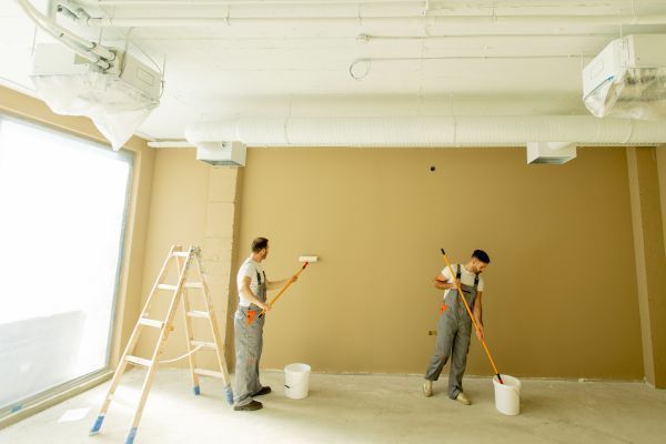 Two painters in a bright room painting a tan wall, with ladders and paint buckets nearby.