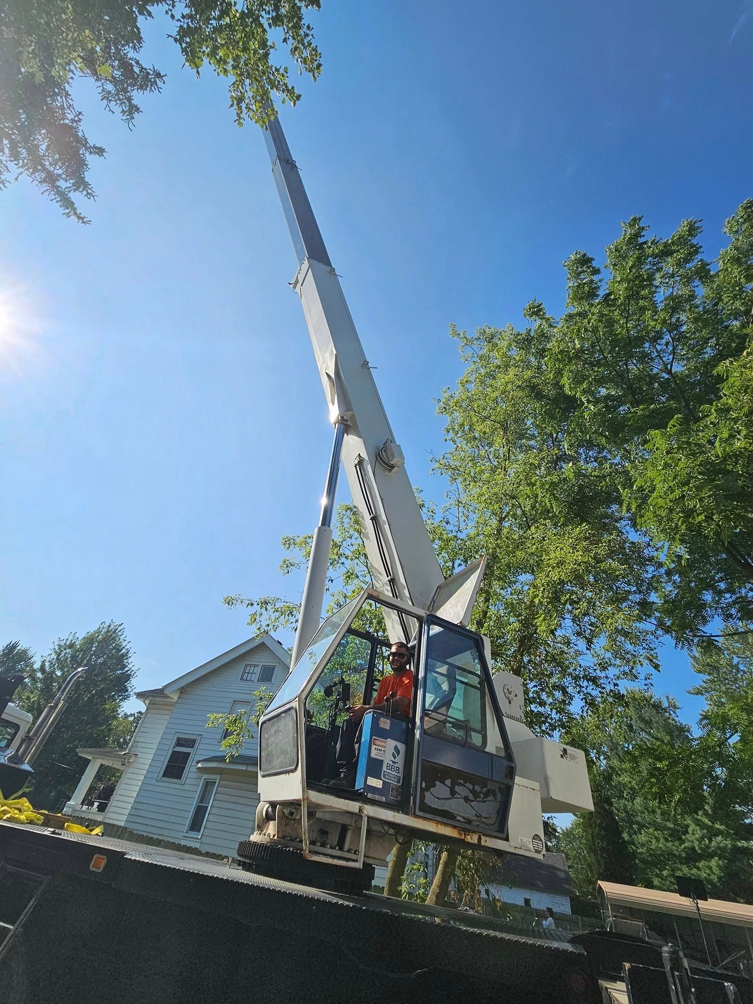 A crane with its boom extended upward sits on a trailer parked next to a house and green trees under a bright blue sky.