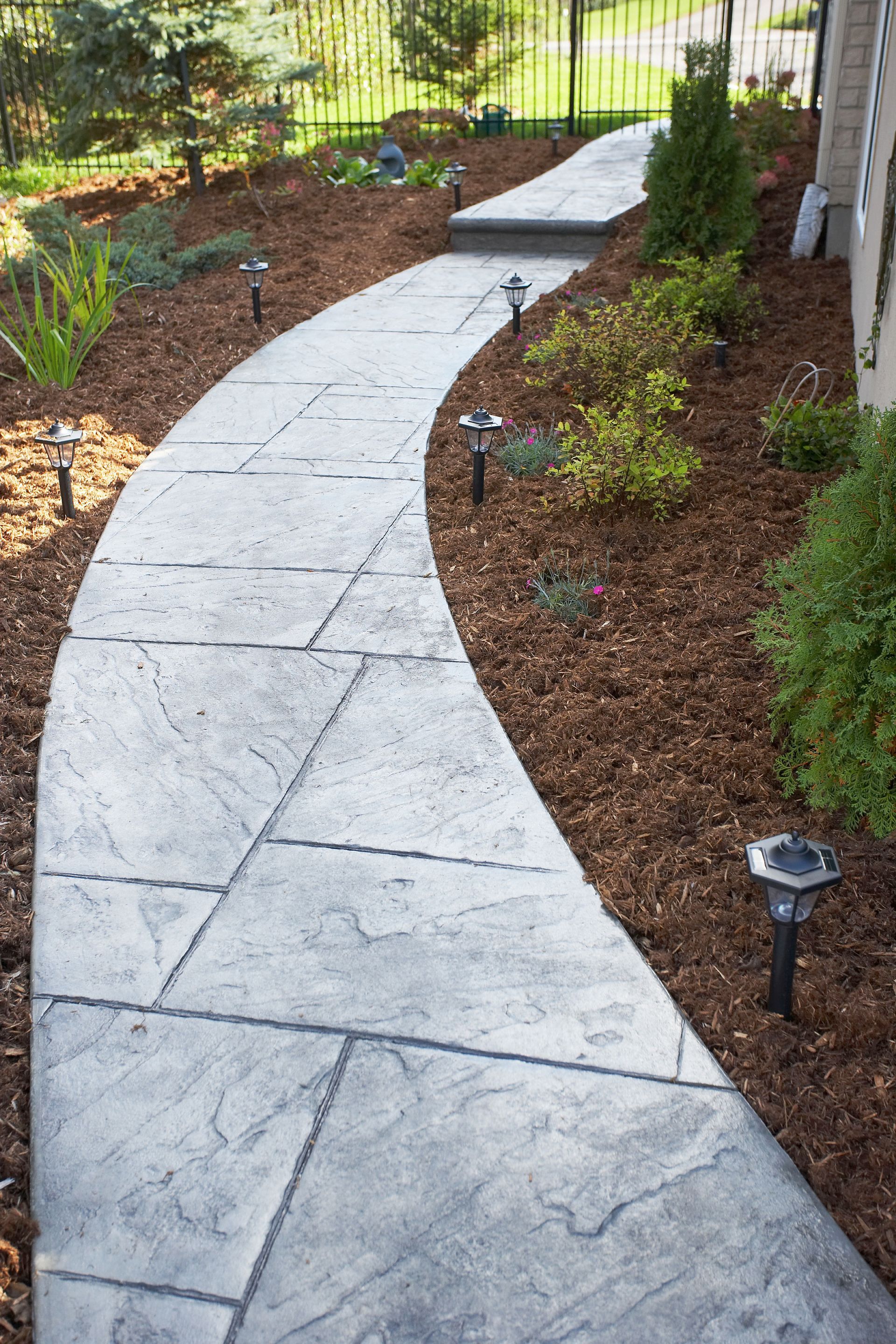 A curved, stamped concrete walkway bordered by mulch and small shrubs with solar lights along a house exterior.