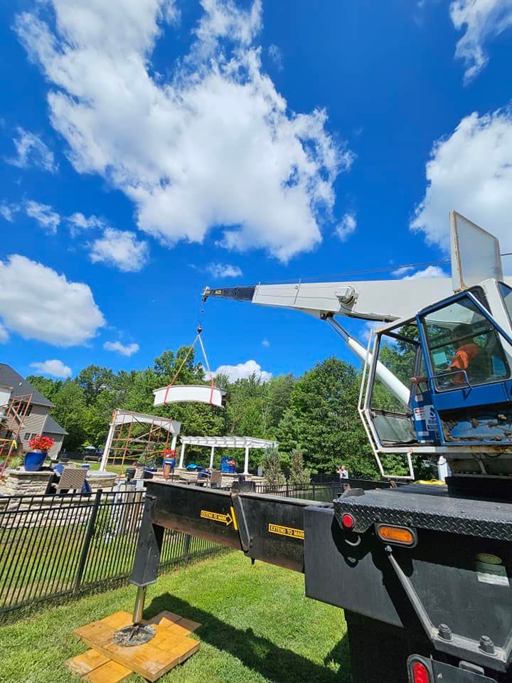 A crane lifting a white, circular hot tub into a residential backyard with trees and blue, cloudy skies in the background.