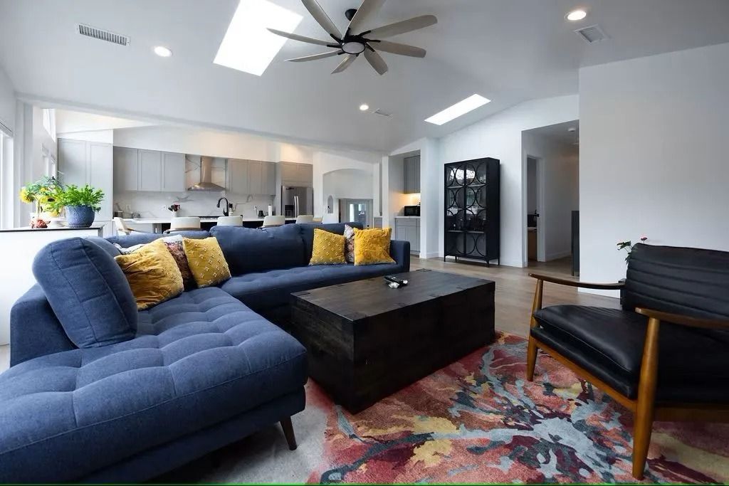 Spacious living room with a blue sectional sofa, coffee table, and colorful rug. Kitchen visible in the background.