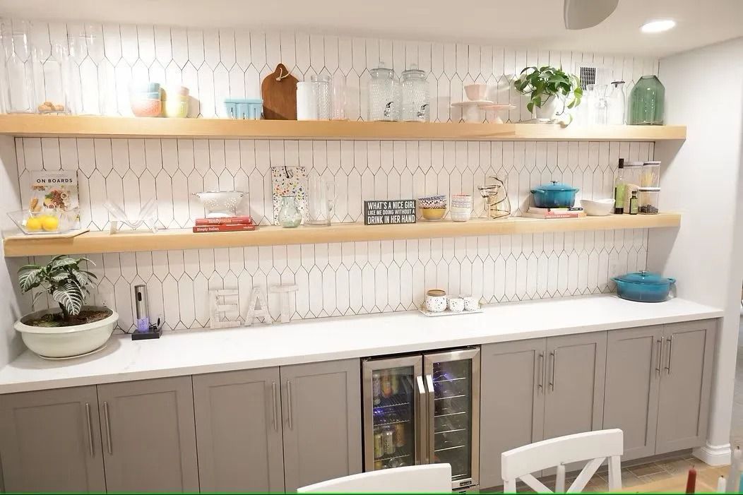 Built-in grey cabinets with a countertop, floating shelves with decor, and a wine fridge. White tiled wall.