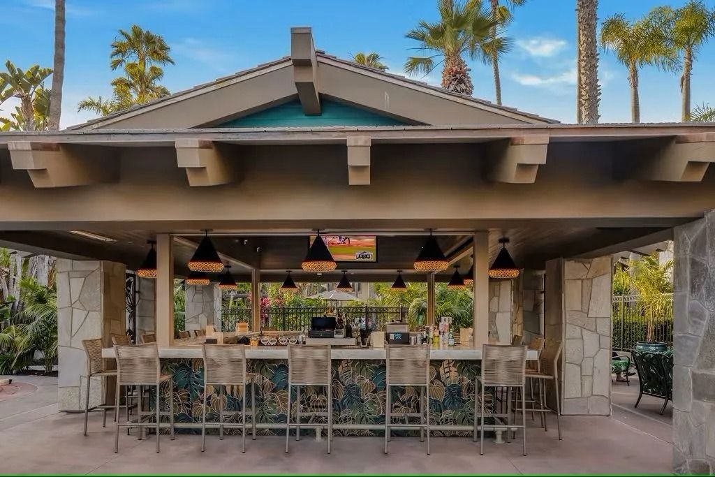 Outdoor bar with stools, under a covered structure with stone columns and palm trees.