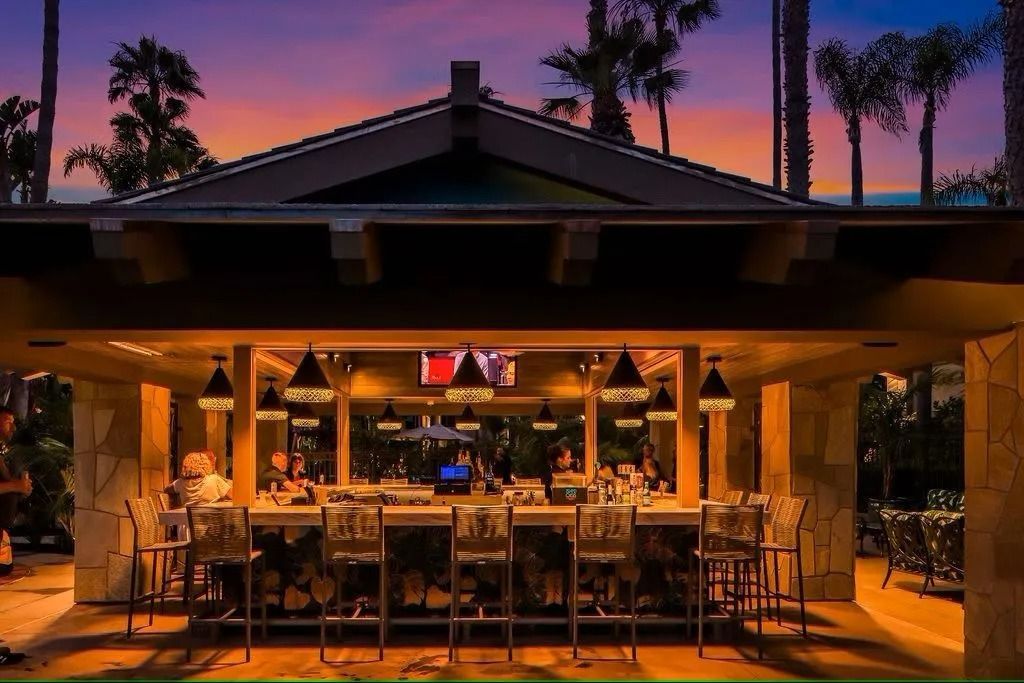Outdoor bar at dusk with lit lamps, palm trees, and sunset sky.