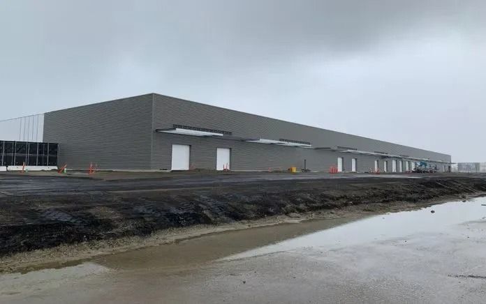 Large gray industrial building under a cloudy sky with loading docks and construction in foreground.