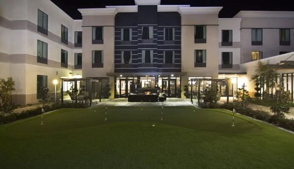 Hotel exterior at night with a putting green in the foreground. Building has multiple stories, lit windows, and neutral colors.